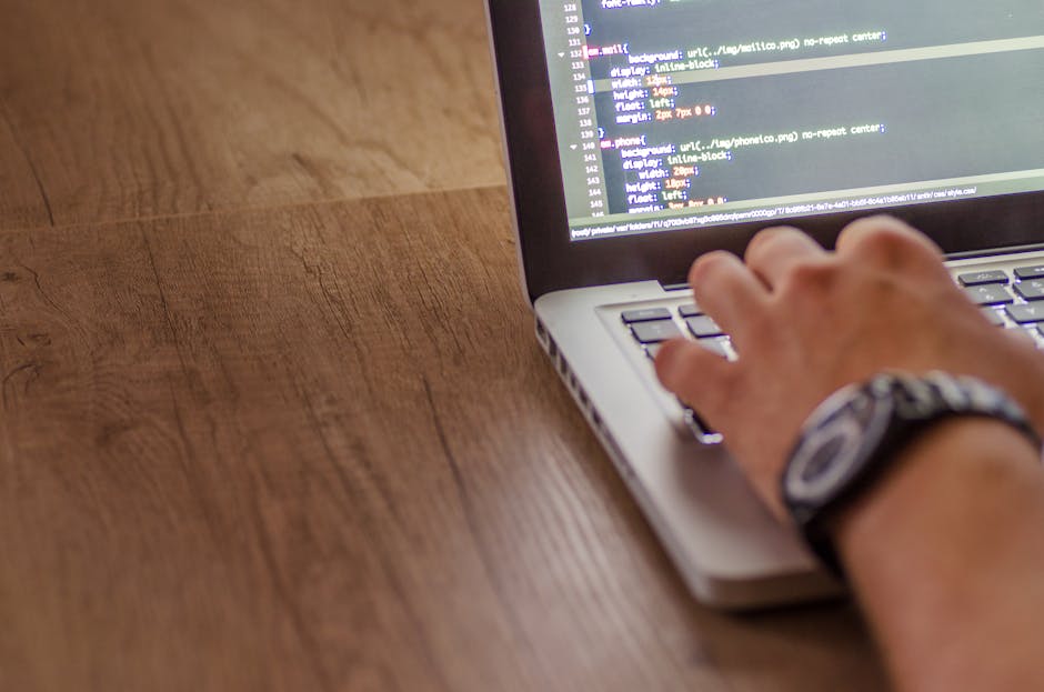 A person's hand with a wristwatch is hovering over a laptop keyboard placed on a wooden surface, with the screen displaying lines of code or text in a notepad, indicating digital work or programming a
