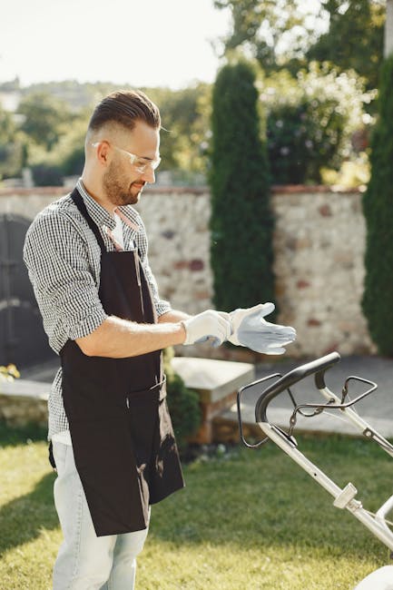 A man with styled hair, glasses, and a trimmed beard, dressed in a checked shirt and dark apron, is standing outdoors in a garden during daytime. He is wearing white gardening gloves and preparing for